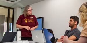 One person standing and two people sitting around a table with laptops having a conversation