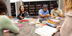 Students studying around a table in a library with books and a tablet.