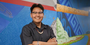 Person standing in front of a colorful mural with arms crossed, wearing glasses and a beaded necklace.