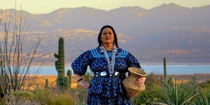 A person in a blue floral dress with silver jewelry stands in a desert landscape with mountains and a body of water in the background.