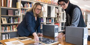 Two people examining photographs and documents on a table in a library.