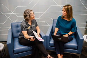 two women seated adjacent to one another in comfy chairs, one with a laptop, having a conversation
