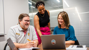 a female standing between a sitting male and sitting female while they look at a laptop
